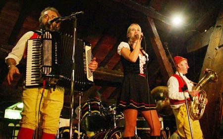 Accordion player, female singer and sousaphone player in red waistcoats on a stage in a timber beamed hall