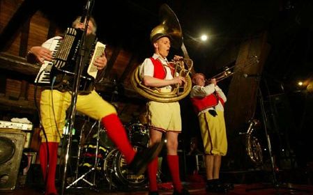 Three musicians with accordion, sousaphone and trombone on stage, the accordion player kicking a leg high