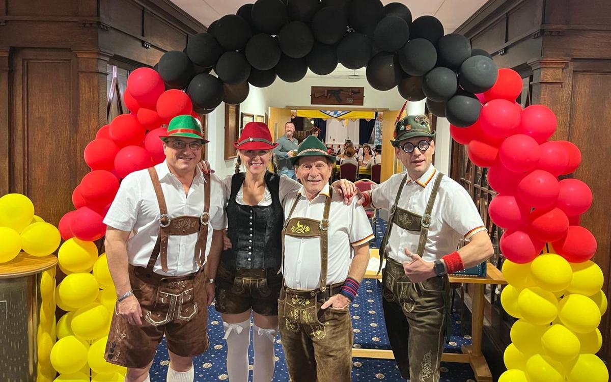 Four band members in lederhosen and Bavarian hats standing under a black, red and yellow balloon arch