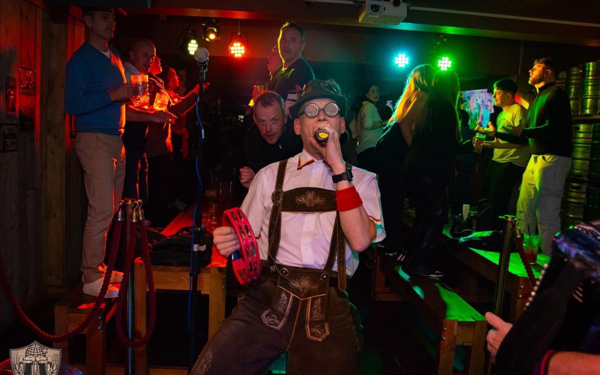 Singer in lederhosen, hat and round glasses holding a tambourine and singing into a microphone among the crowd