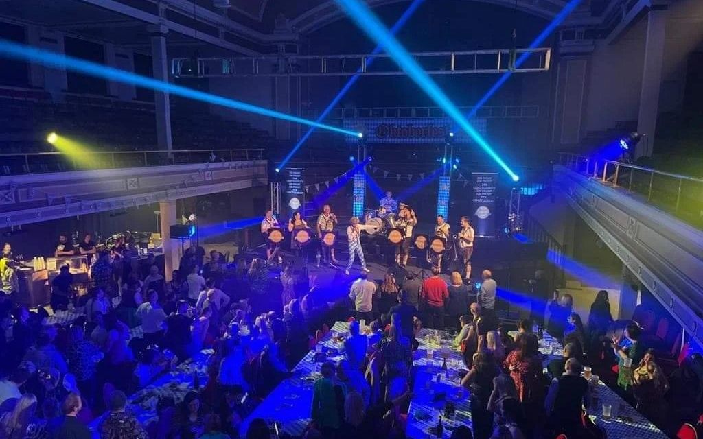 The band on stage in a large hall lit by blue laser beams, with a seated crowd at long tables