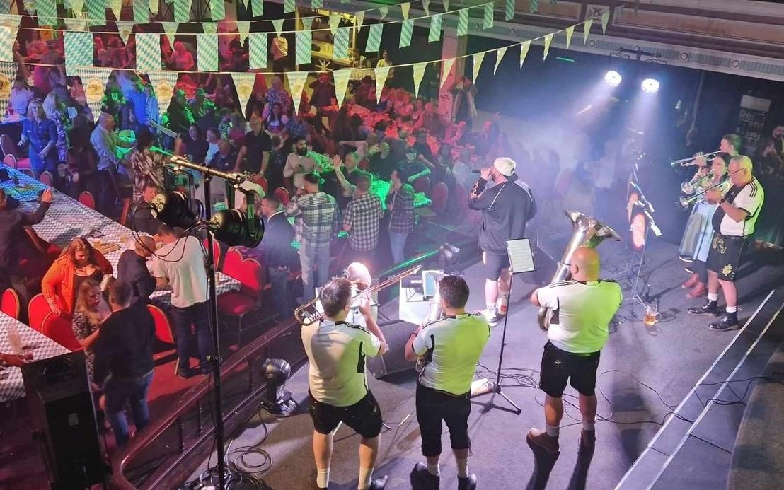 The band seen from above playing brass on stage in a hall hung with blue and white bunting, with a crowd at long tables