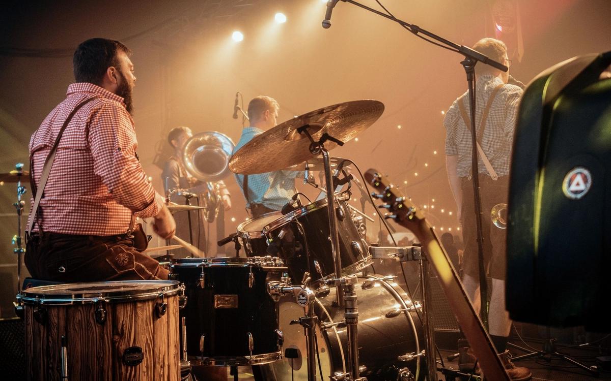 Band in lederhosen on a large stage under warm lights, seen from the side, playing to a packed festival tent
