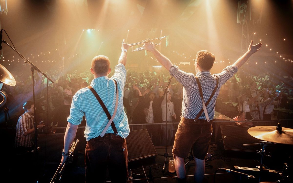 Two players in lederhosen seen from behind with arms raised on stage, facing a large crowd under strings of lights