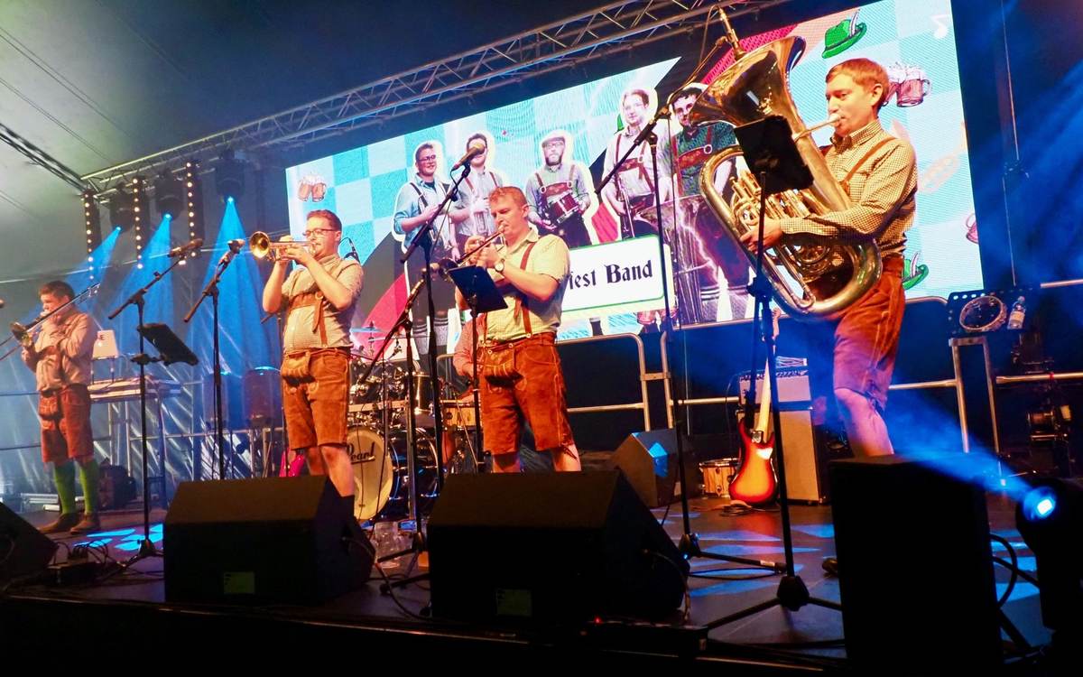 Band in lederhosen performing on a stage with a tuba, trumpets and trombone in front of a bright video screen