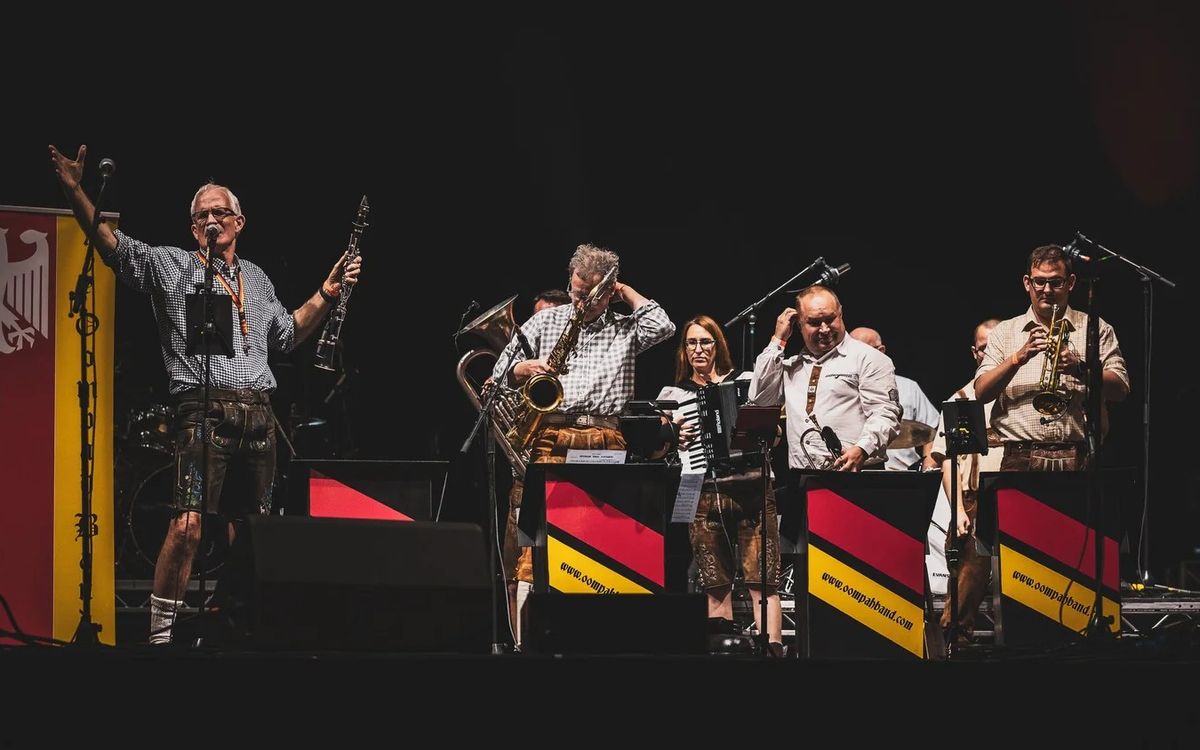 Band on a dark stage in lederhosen and checked shirts, the front man with arms raised holding a clarinet, beside German flag music stands