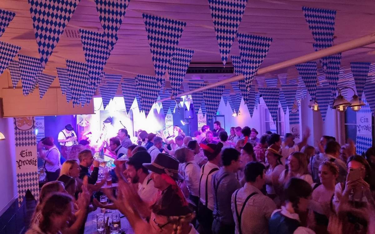 Packed room of guests in Bavarian hats at long tables under blue and white bunting, with the band on stage at the far end